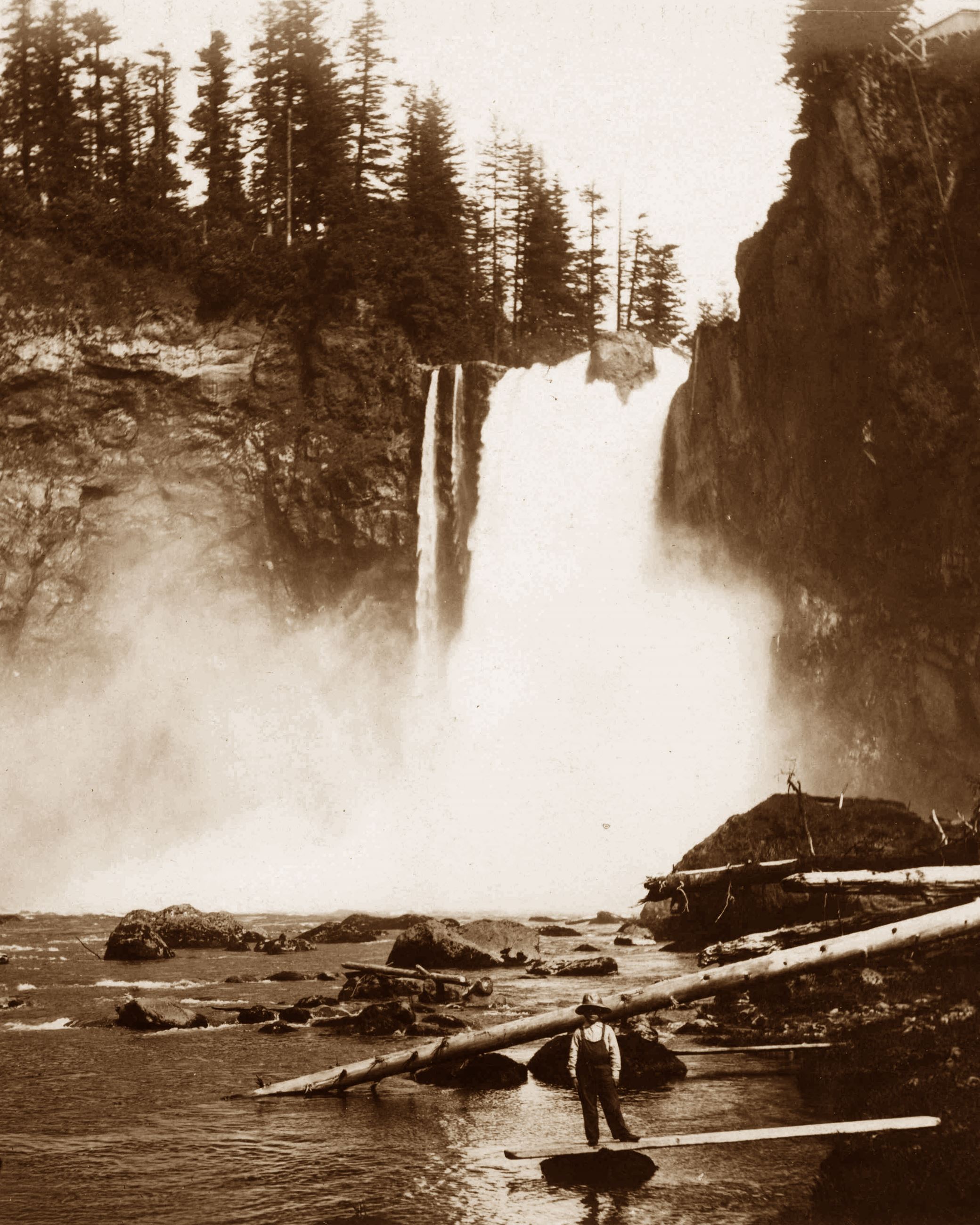 This is a black and white photo of a man standing at the bottom of Snoqualmie Falls around 1900.